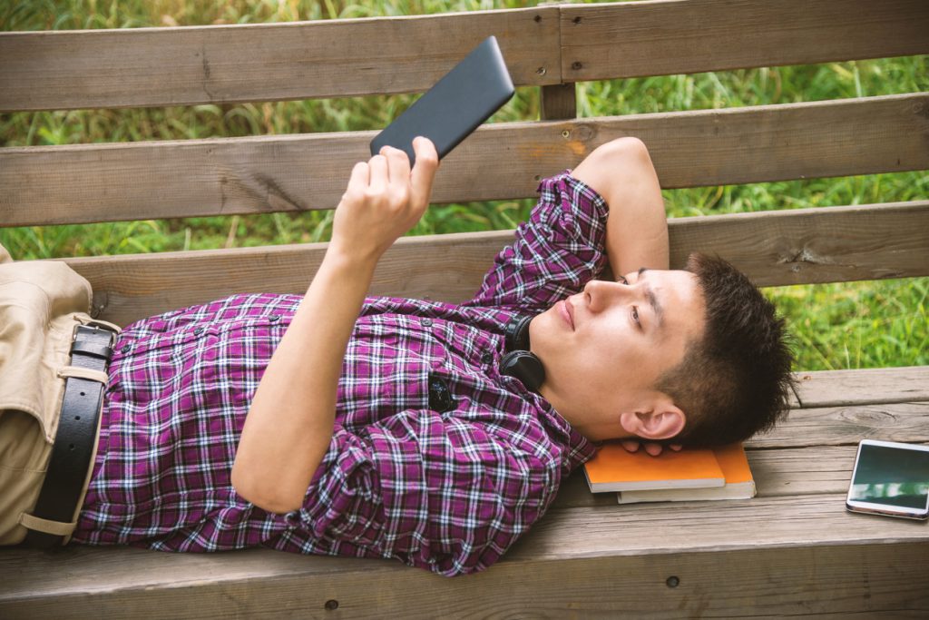 Handsome Asian man reading digital tablet lying on bench